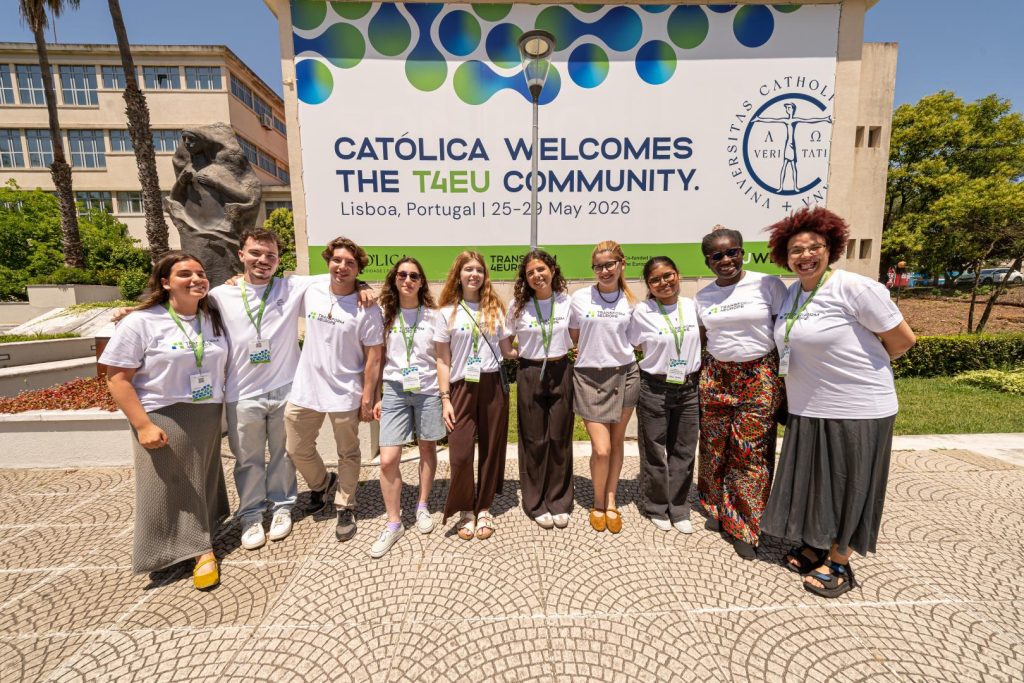 A diverse group of ten smiling young adults stand in front of a large banner that reads, Católica welcomes the T4EU Community. Lisboa, Portugal, 25-29 May 2026. They wear event badges and stand outdoors on a sunny day.