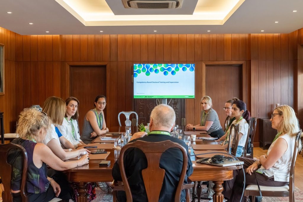 A group of people are seated around a conference table in a wood-paneled meeting room, facing a screen displaying a presentation titled Compassionate About Dementia Training and Awareness.