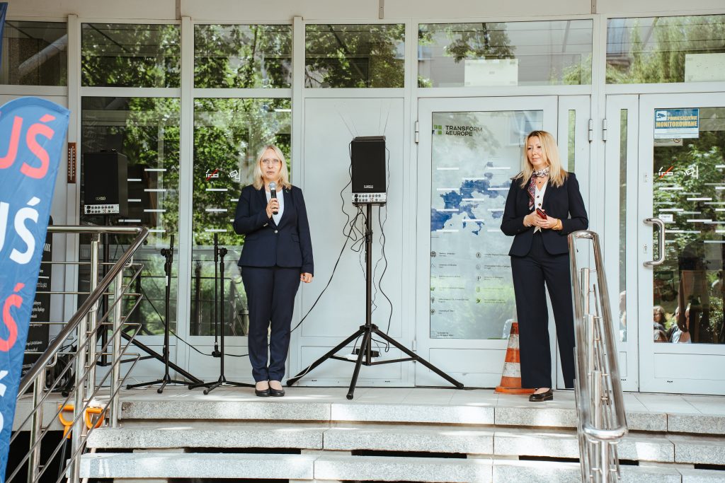 Two women in business suits stand on steps outside a building, each holding a microphone. Behind them are glass doors, speakers, a map, and an orange traffic cone. Green trees are reflected in the glass.