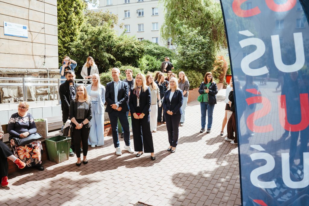 A group of people, dressed mostly in business attire, stand outdoors on a brick walkway near a building, listening attentively. A blue banner with red and white letters is partially visible on the right. Trees and greenery surround the scene.