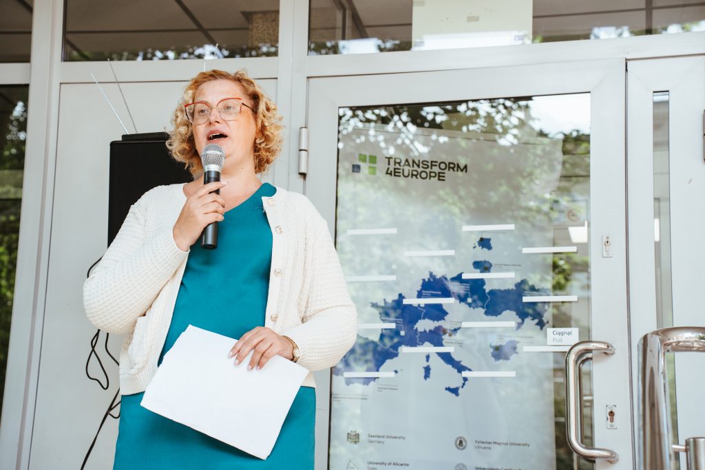 A woman with curly blonde hair, glasses, and a teal dress, holding a microphone and papers, speaks in front of a glass door with a Transform 4 Europe map and logo displayed behind her.