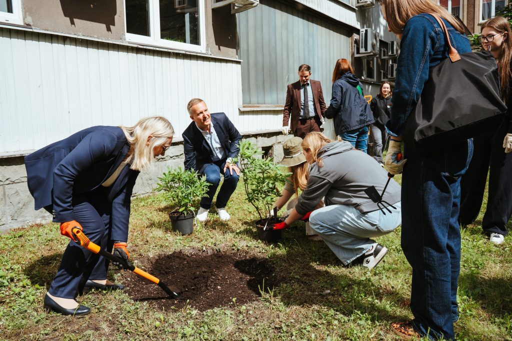 A group of people are gathered outside a building, planting small bushes in the grass. One woman is digging with a small shovel, while others watch or assist with the planting. The weather appears to be sunny.