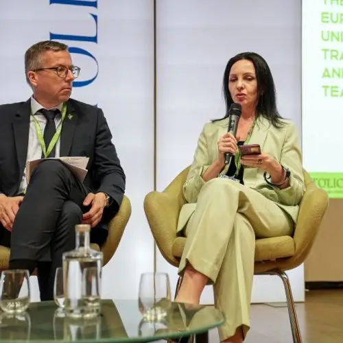 Two people sit on stage in chairs at a panel discussion. The woman on the right is speaking into a microphone and holding a phone. The man on the left listens attentively. A screen with event text is visible behind them.