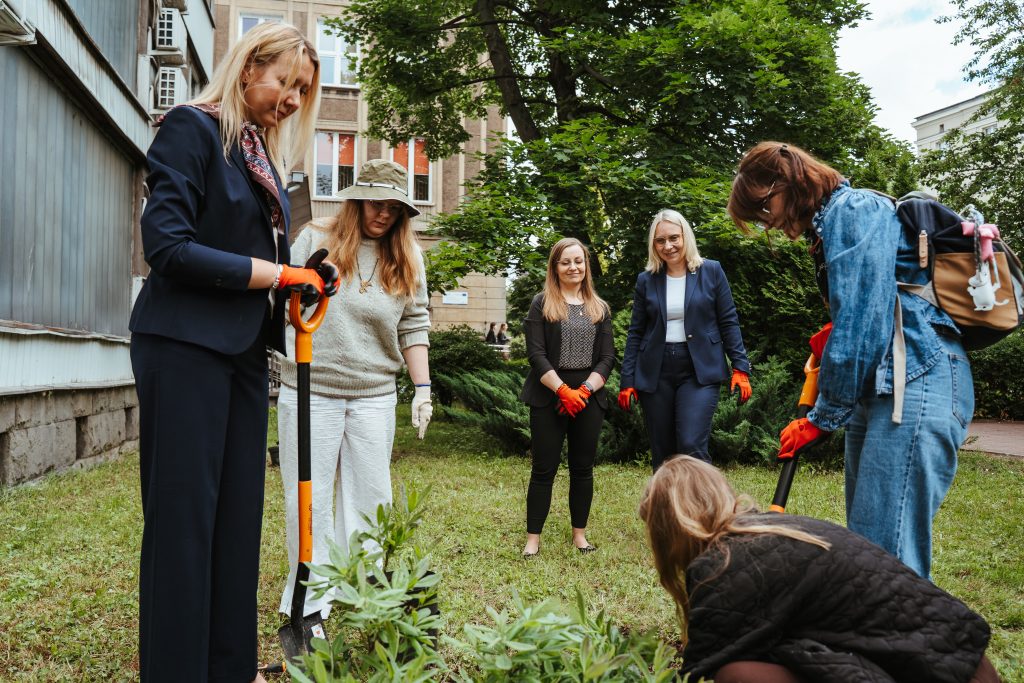 Five women stand on a grassy area outside a building, some holding shovels and wearing gloves, appearing to plant or tend to greenery. Trees and bushes are in the background.