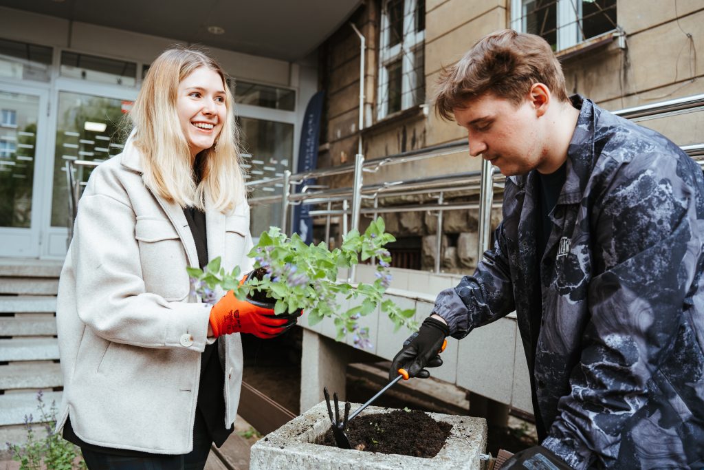 Two people gardening together outdoors; a smiling woman in a light jacket and red gloves holds a plant, while a man in dark clothes plants something in a concrete pot filled with soil.