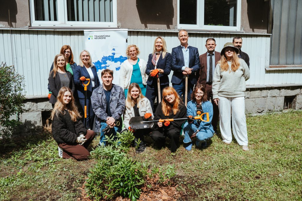A group of people, some holding shovels, pose together outdoors in front of a building and a sign. They are standing and kneeling on the grass near a small shrub, suggesting a community or environmental activity.