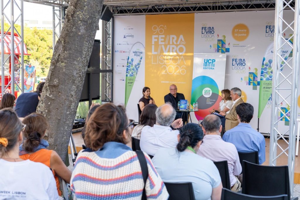 A seated audience listens to a panel of four speakers on stage at the 96th Lisbon Book Fair 2026, under a tent with colorful banners and a tree in the foreground.