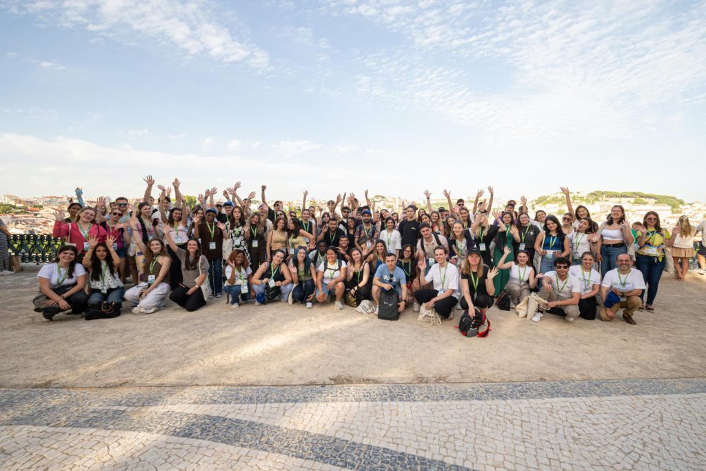 A large group of people pose together outdoors on a sunny day, many smiling and raising their arms. The background shows a scenic cityscape and blue sky with scattered clouds.