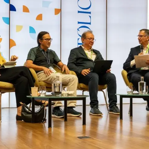 Four people sit on stage in armchairs, having a panel discussion. They hold papers and laptops, with water glasses on tables in front. A screen behind them displays “AI ENABLED GLOBAL ACADEMIC COLLABORATION.”.