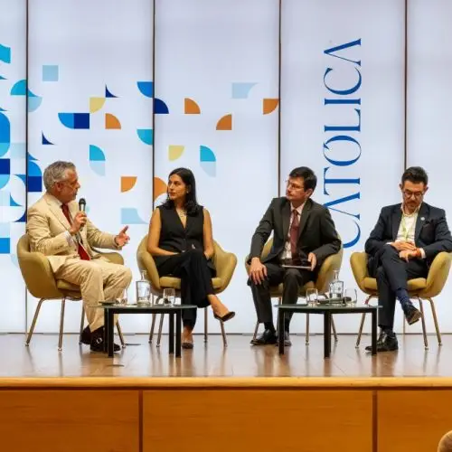 Four people in formal attire sit on stage in armchairs, engaged in a panel discussion. Behind them is a backdrop with abstract patterns and the word CATOLICA. Glasses of water sit on small tables beside them.