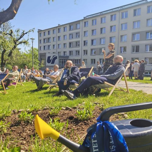 People sit on deck chairs and grass in front of a modern, multi-story building on a sunny day. A woman stands speaking, and a watering can with a blue ribbon is in the foreground. Trees and greenery are visible.