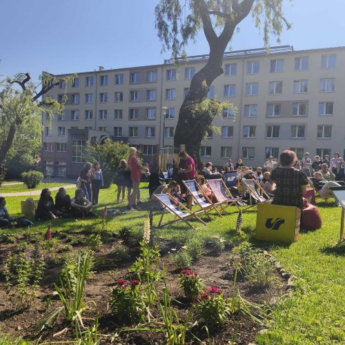 People gather outdoors on a sunny day, sitting on deck chairs and grass near a small garden with flowers and plants. In the background, there is a large apartment building and a tall tree.
