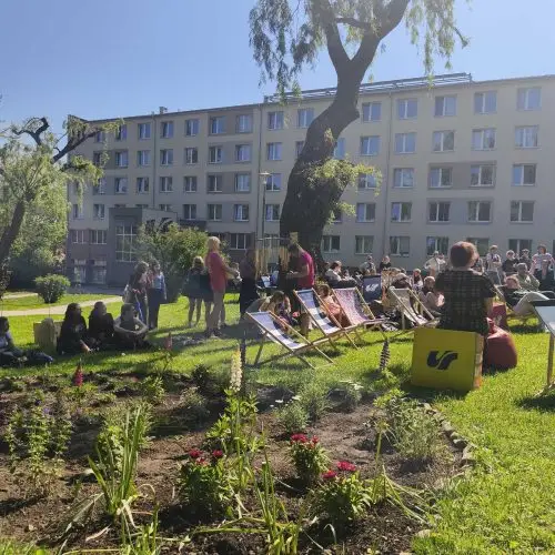 People gather outdoors on a sunny day, sitting on deck chairs and grass near a small garden with flowers and plants. In the background, there is a large apartment building and a tall tree.