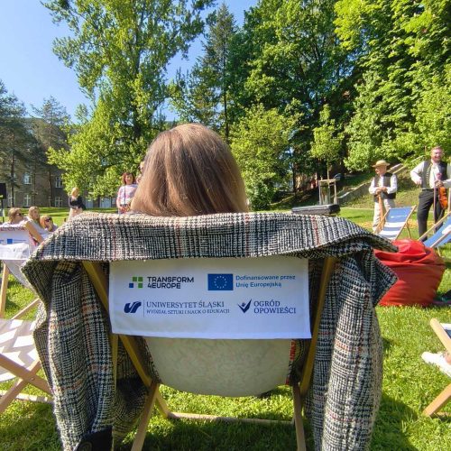 A person sits on a deck chair outdoors on a sunny day, with a jacket draped over the chair. The chair has logos and text, and several people are gathered in the background on a grassy area surrounded by trees.