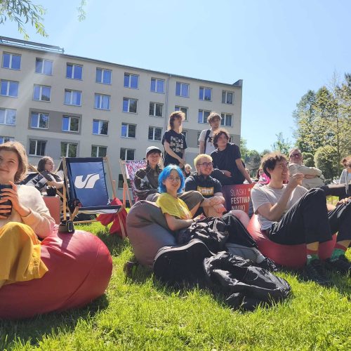 A group of young people relax on bean bags and deck chairs on a grassy lawn in front of a modern building, enjoying a sunny day at an outdoor event or festival.