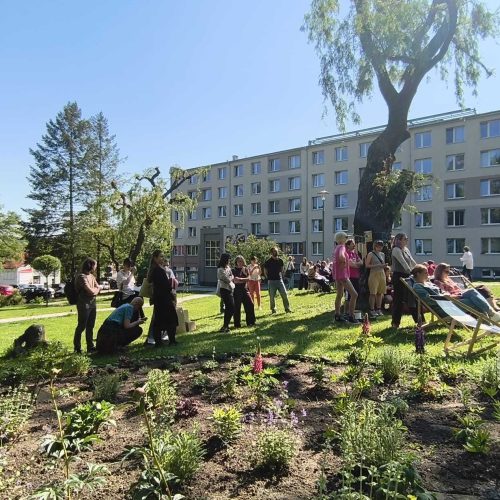 People gather and socialize in a sunny park with flower beds, lawn chairs, and bean bags. Apartment buildings and trees are visible in the background under a clear blue sky.
