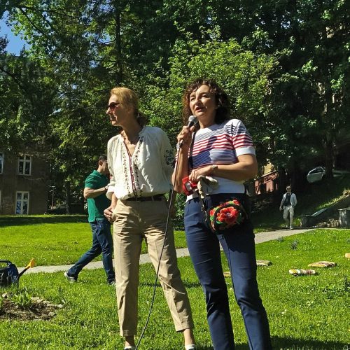 Two women stand on green grass in a sunny park, one holding a microphone and speaking, the other smiling. People and trees are visible in the background under a clear blue sky.