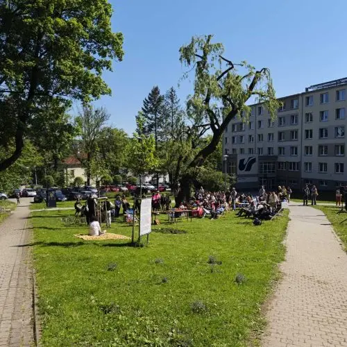 A group of people gathers on a grassy area near a large tree and a modern building on a sunny day. Some sit on blankets, while others stand or sit on benches. The scene is bordered by paved walkways and trees.