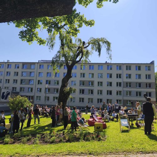 A large group of people gathers on a sunny lawn in front of a multi-story building, enjoying outdoor activities and socializing under the shade of trees. Tables and chairs are set up across the grassy area.