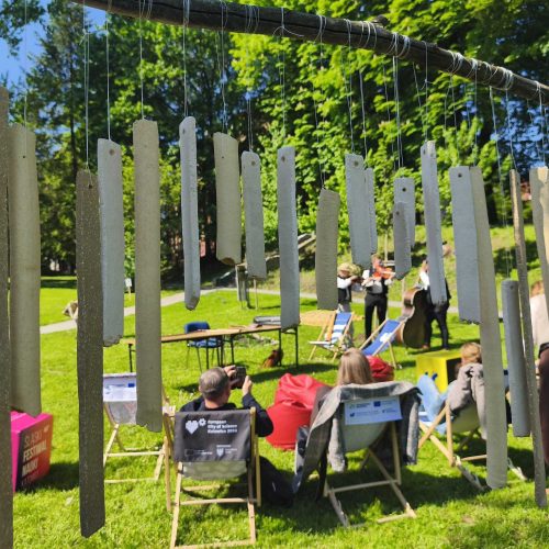 People sit on deck chairs and colorful beanbags on a sunny lawn, watching performers in black attire. Cardboard chimes hang in the foreground. Tall trees and a building are visible in the background.