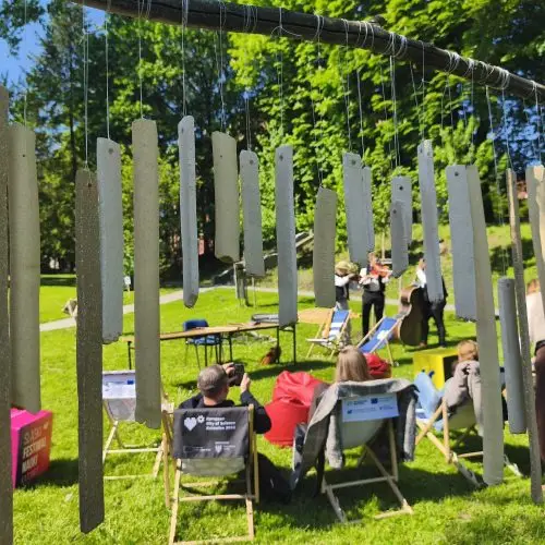People sit on deck chairs and colorful beanbags on a sunny lawn, watching performers in black attire. Cardboard chimes hang in the foreground. Tall trees and a building are visible in the background.