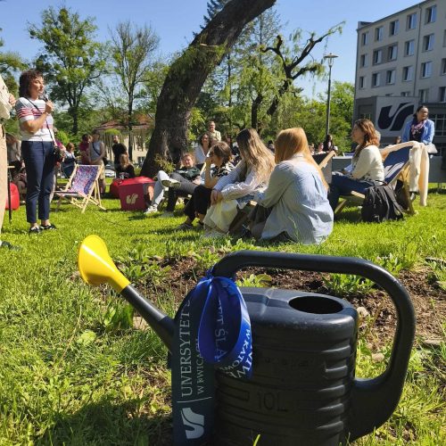 A black watering can with a blue ribbon sits on green grass in the foreground, while a group of people sit and talk in a sunny park, with trees and a multi-story building in the background.