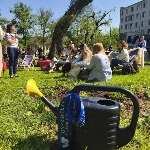A black watering can with a blue ribbon sits on green grass in the foreground, while a group of people sit and talk in a sunny park, with trees and a multi-story building in the background.