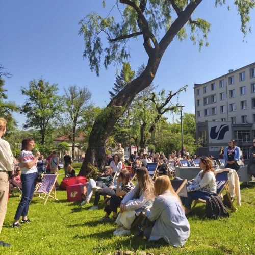 A group of people are gathered outdoors on a sunny day, sitting on the grass and lounge chairs under a large tree in front of a modern building. A woman is speaking to the audience while others listen attentively.
