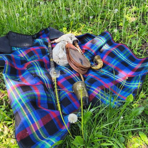 A blue, green, and red plaid blanket lies on grass, with bagpipes and accessories resting on top. Dandelions and green plants surround the blanket in a sunny outdoor setting.