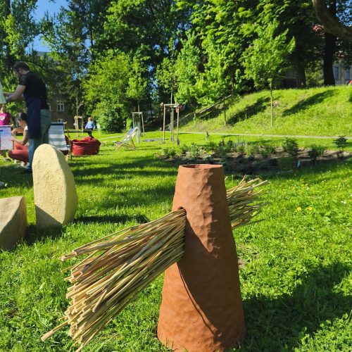 A brown cone-shaped object with bundles of straw sticking out from its sides stands on green grass in a sunlit park, with trees, people, and playground equipment in the background.