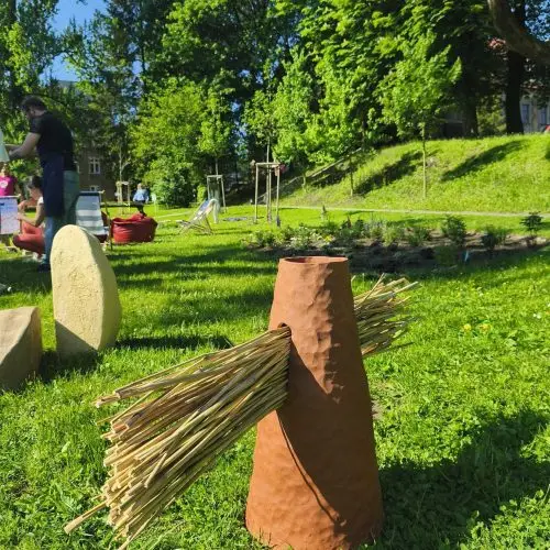 A brown cone-shaped object with bundles of straw sticking out from its sides stands on green grass in a sunlit park, with trees, people, and playground equipment in the background.
