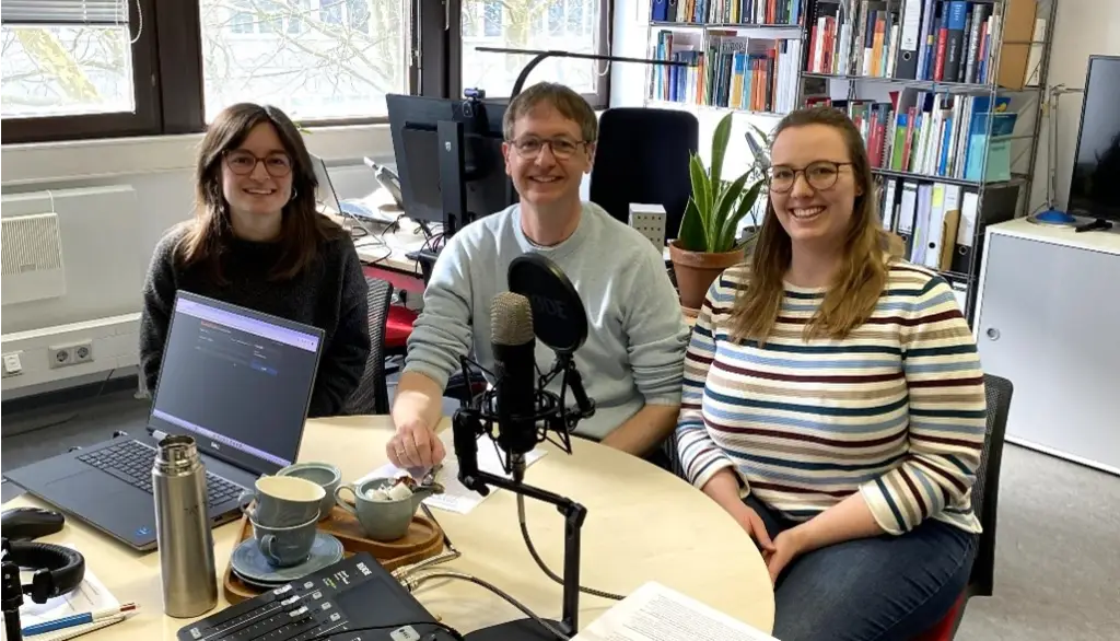 Three people sit smiling around a round table with microphones, a laptop, audio equipment, cups, and a plant, in a bright office with bookshelves and large windows in the background.