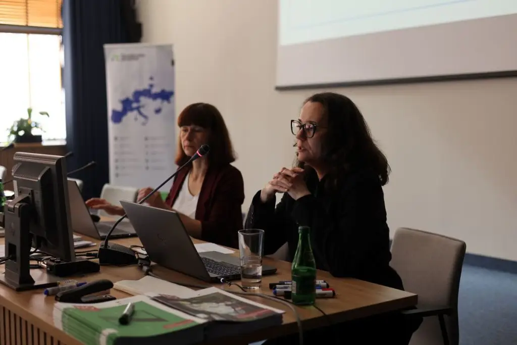 Two women sit at a conference table with laptops and papers, speaking into microphones. A presentation is projected on the screen behind them. Bottled water and office supplies are on the table.