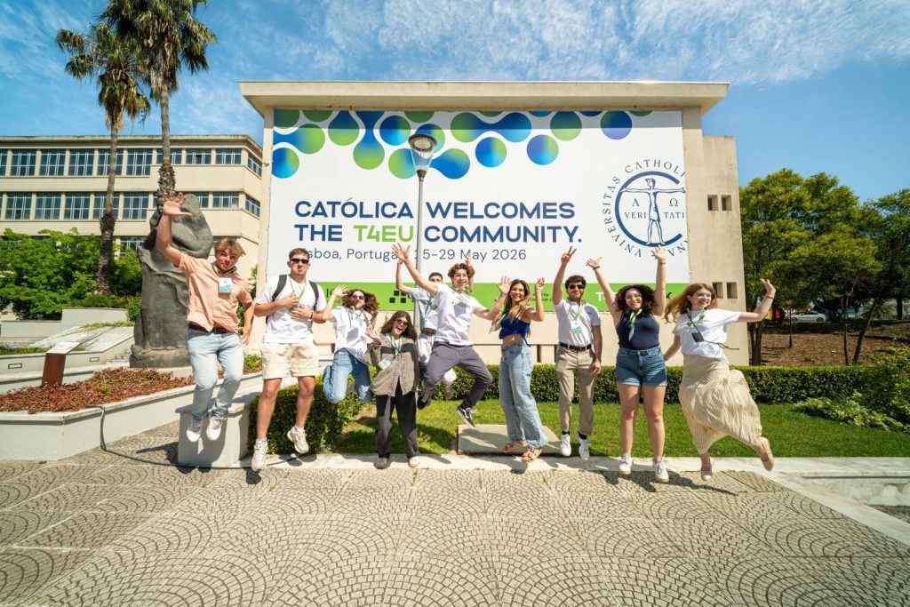 A group of young people jump and pose in front of a large sign that reads “Católica welcomes the T4EU community. Lisboa, Portugal, 25-29 May 2026.” The background includes buildings, trees, and a bright blue sky.