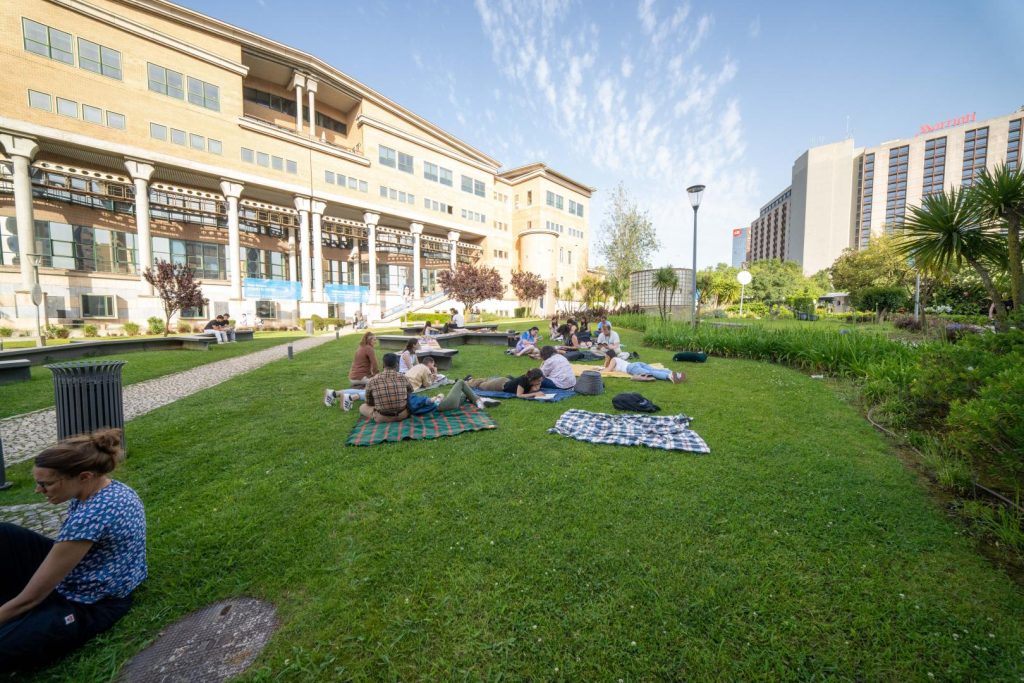 People relax on blankets on a grassy lawn near a modern building on a sunny day, with another tall building and lush greenery in the background.