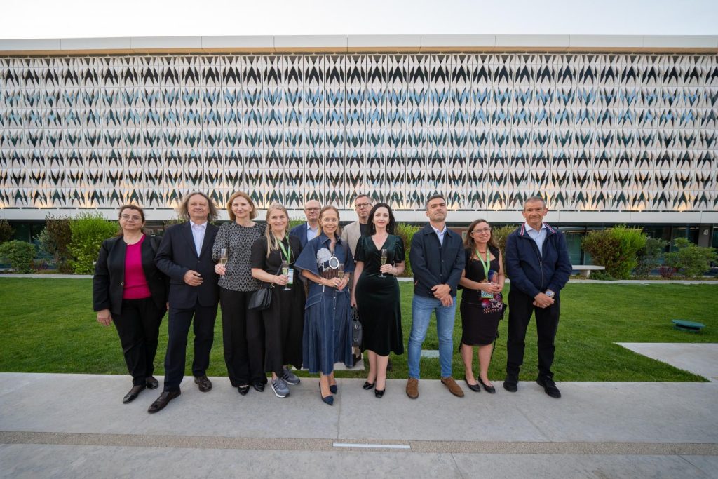 A group of ten people dressed in formal attire stand in a row outdoors, each holding trophies or awards, with a modern building featuring a geometric facade in the background.