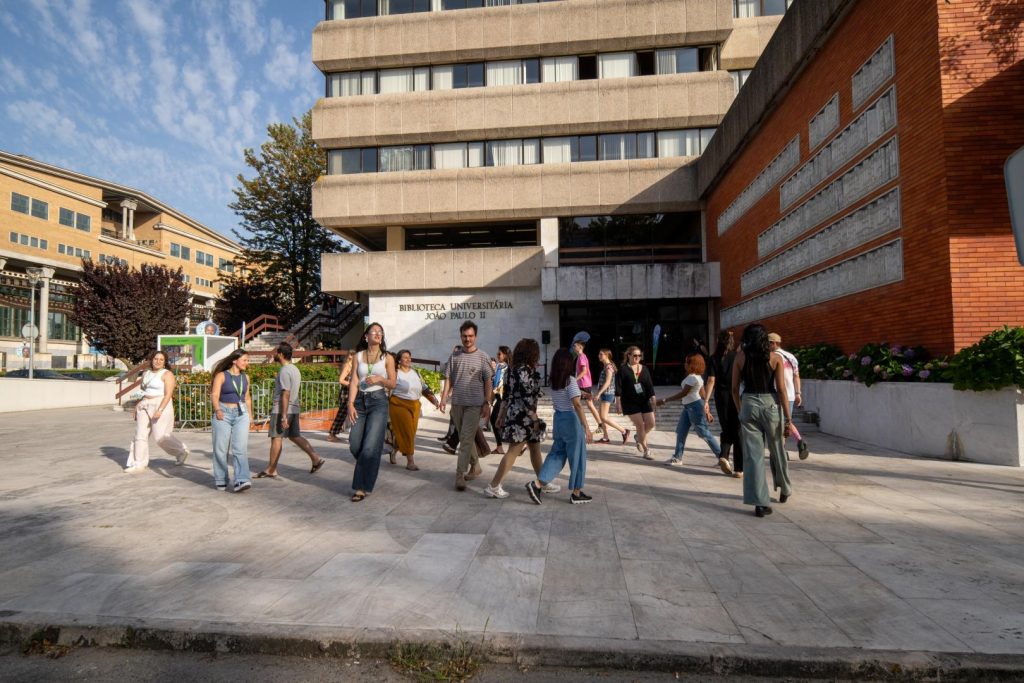 A group of people, mostly young adults, walk in front of a modern university library building under a blue sky with scattered clouds. Some are chatting while others walk alone.