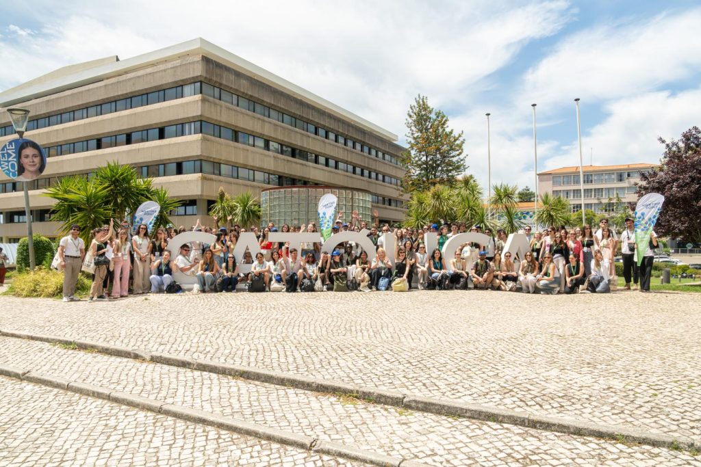A large group of people pose outdoors in front of big white letters spelling CATOLICA, with modern buildings, palm trees, and a partly cloudy sky in the background.