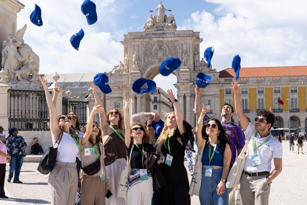 A group of people joyfully toss blue caps into the air in a sunny plaza, with a grand arch and historic buildings in the background. Most are smiling and wearing lanyards, suggesting a celebratory or group event.