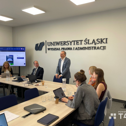 A group of people sit and work on laptops around a conference table in a meeting room at Uniwersytet Śląski, Wydział Prawa i Administracji. One man stands and addresses the group; a screen displays a website.