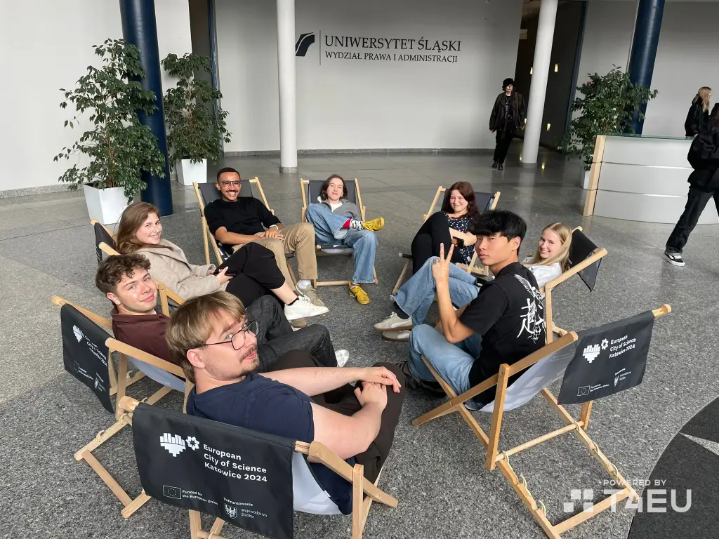 A group of eight young adults sit in a circle on folding chairs indoors, smiling and chatting. Behind them is a sign for the University of Silesia, Faculty of Law and Administration.