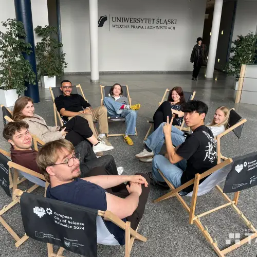 A group of eight young adults sit in a circle on folding chairs indoors, smiling and chatting. Behind them is a sign for the University of Silesia, Faculty of Law and Administration.