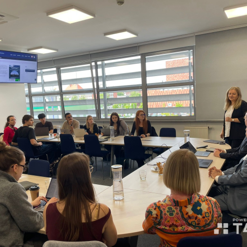 A group of people sit around tables in a modern conference room, listening to a woman standing and speaking. Laptops and papers are on the tables, and presentation screens display project slides. The logo Powered by T4EU is visible.