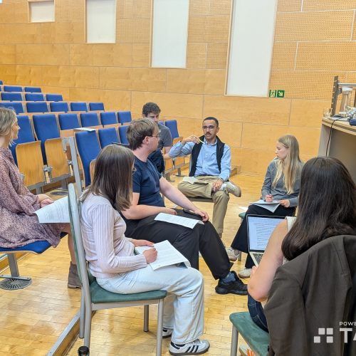 A small group of students sits in a circle in a lecture hall, engaged in discussion with notebooks and laptops. Most seats are empty, and a lecturers podium is visible at the front.