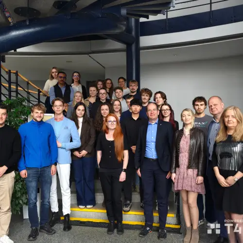 A group of people poses for a photo on a staircase indoors, with some standing on steps and others at the bottom. They are dressed in smart casual attire. A spiral staircase and greenery are visible in the background.