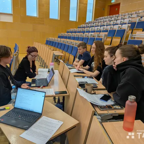 Six students sit in a lecture hall, working together at desks with laptops, papers, and drinks. They appear focused, discussing or collaborating. The room has wooden walls and rows of empty blue chairs.