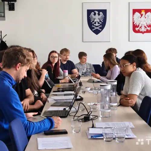 A group of people sit around a conference table with laptops and papers, engaged in discussion. Two Polish coat of arms emblems hang on the wall in the background.