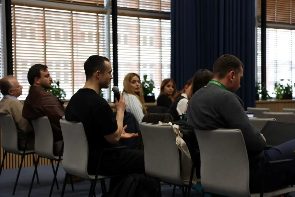 A man in a black shirt speaks into a microphone while seated among other attendees in a conference room with large windows and blue curtains.