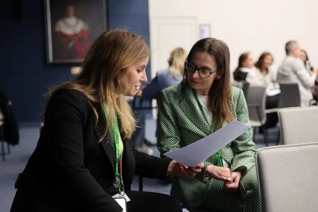 Two women sit and discuss a document in a conference room setting, with other people blurred in the background. One woman wears a black blazer, the other a green checkered suit. Both have conference badges.