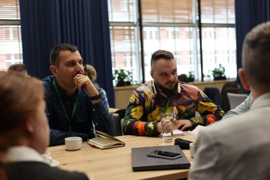 Four people sit around a table in a well-lit room, engaged in discussion. Two men with lanyards are in focus, with notebooks, a phone, and drinks on the table. Large windows with blinds are in the background.